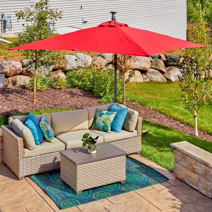 A red umbrella shades a cushioned outdoor sofa and coffee table on a patterned rug, surrounded by greenery and stone landscaping in a sunny yard.