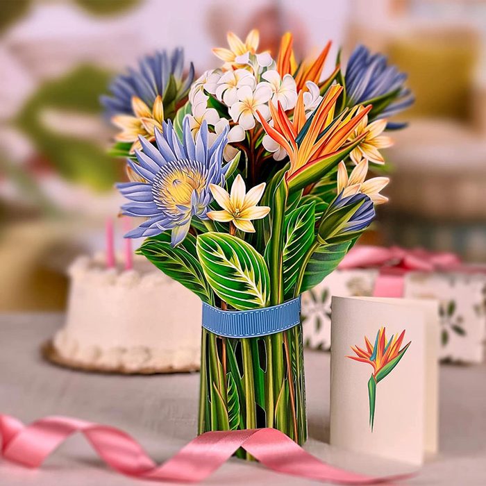 A vibrant bouquet of colorful flowers stands in a decorative vase, surrounded by a cake and gift boxes with ribbons on a table.
