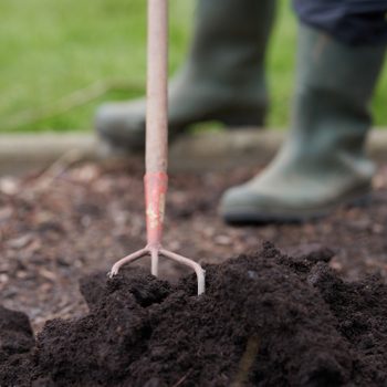 A gardening fork digs into dark, rich soil while a person in rubber boots stands nearby on a grassy background.