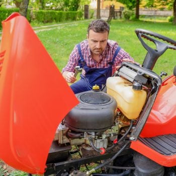 A person inspects a small engine under the hood of a red riding lawn mower, surrounded by green grass and trees in a backyard.