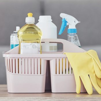A pink cleaning basket holds various cleaning supplies, including bottles and gloves, positioned on a wooden surface against a soft, neutral background.
