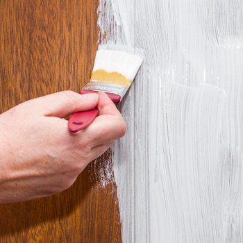 A hand paints a section of wood with a brush, applying white paint. The contrasting wood grain is visible beside the painted area.