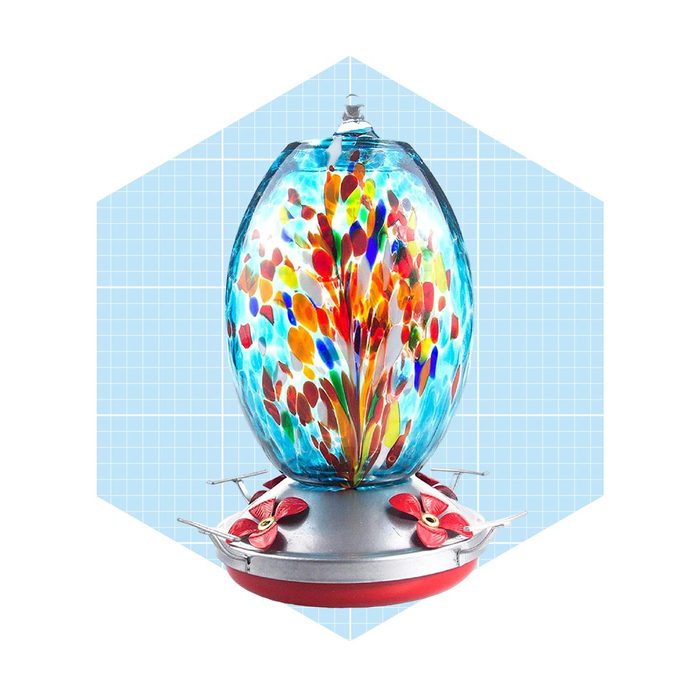 A colorful glass hummingbird feeder stands centrally, filled with nectar. It is supported by a metal base, positioned against a light blue geometric background.