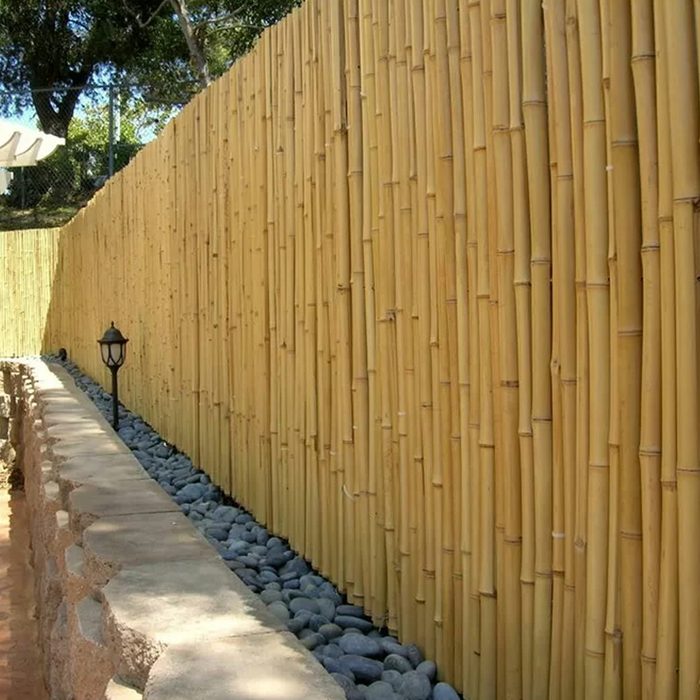 A tall bamboo fence stands alongside a stone path. Smooth stones line the base, with a lamp post casting gentle light in the area.
