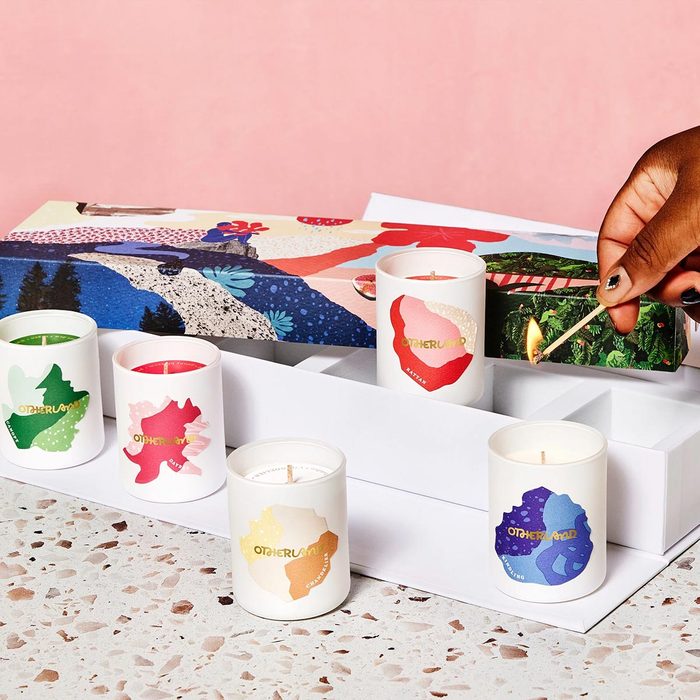 A hand lights a match, igniting a candle among several decorative candles in a white box with an artistic, vibrant background.
