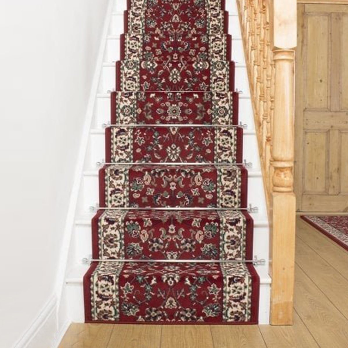 A decorative red carpet runner is laid on white stairs, providing a contrast to the wooden rail and floor in a well-lit interior space.