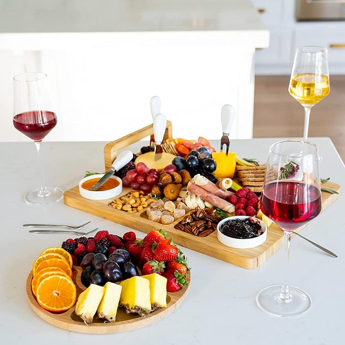A wooden platter displays assorted cheeses, fruits, nuts, and meats, accompanied by two glasses of colored wine, set on a light countertop.