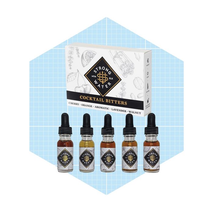 Five glass bottles of cocktail bitters, labeled in various flavors, are arranged in front of a decorative box, set against a light blue geometric background.