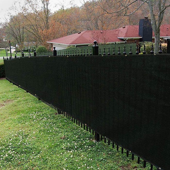 A tall, black privacy fence stands along a grassy area, offering seclusion in a suburban setting with houses and trees in the background.
