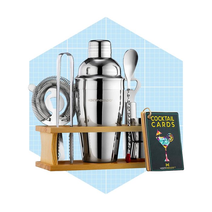 A cocktail shaker set, including a shaker, strainer, spoon, and cocktail cards, is displayed on a wooden stand against a light blue grid background.