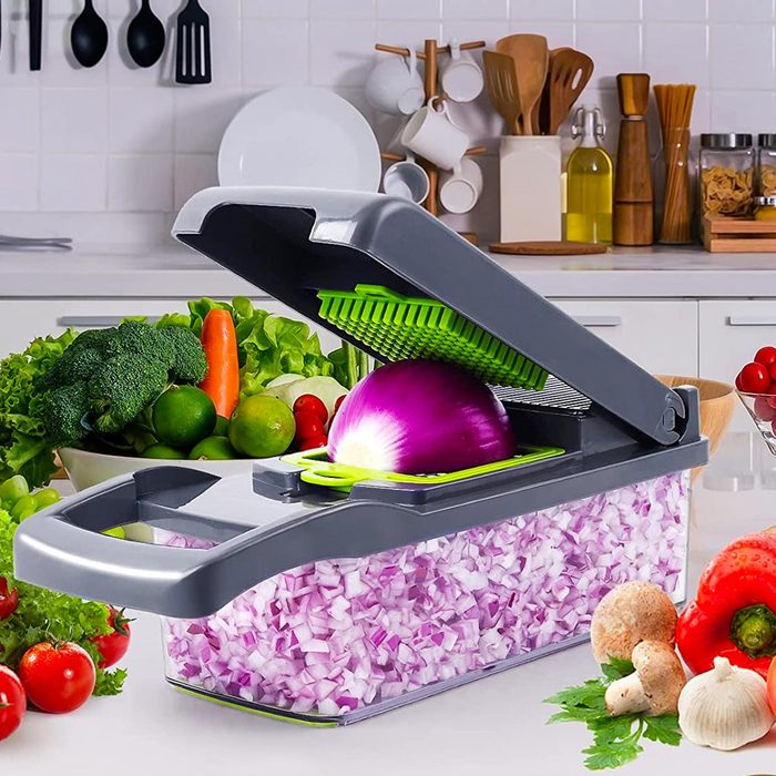 A vegetable chopper with an open lid holds a halved onion above a container filled with diced onions, surrounded by fresh produce on a kitchen countertop.