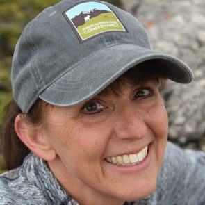 A woman wears a cap with a park logo, smiling warmly while posed outdoors, surrounded by natural scenery and rocky elements.