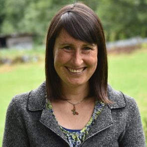 A woman with dark brown hair and freckles smiles, wearing a gray blazer over a floral top, standing in a green, outdoor setting.