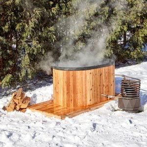 A wooden hot tub steams in a snowy outdoor setting, accompanied by a stacked firewood pile and a metal heating apparatus beside it.