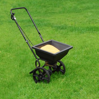 A black wheeled spreader filled with yellow material stands on a lush green lawn, ready for use in garden maintenance.