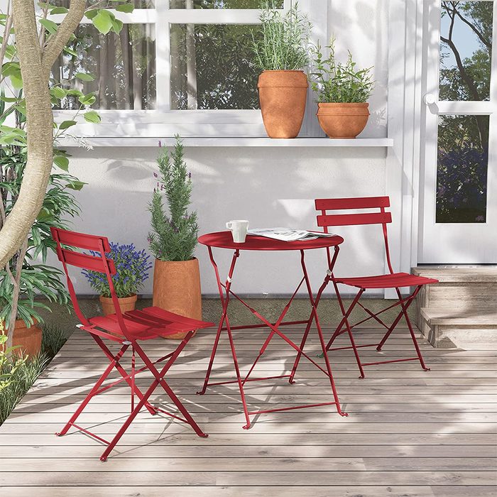 Two red folding chairs and a round table sit on a wooden porch surrounded by potted plants and a door, inviting relaxation in a sunny spot.