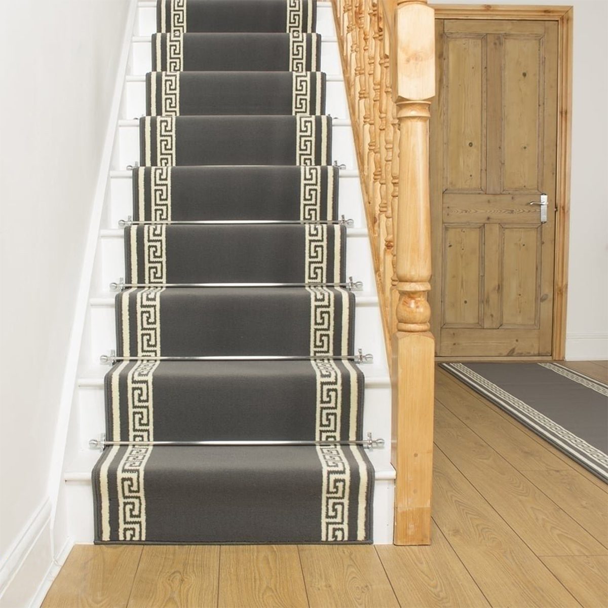 A staircase with dark carpet runners features a decorative pattern, flanked by wooden banisters, leading to a wooden door in a bright room.