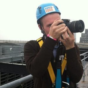 A person wearing a blue helmet and harness is taking a photo with a camera, standing on a rooftop structure in a cloudy environment.
