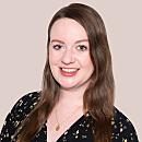 A woman with long brown hair smiles while wearing a black patterned blouse. The background is a plain light color, emphasizing her cheerful expression.