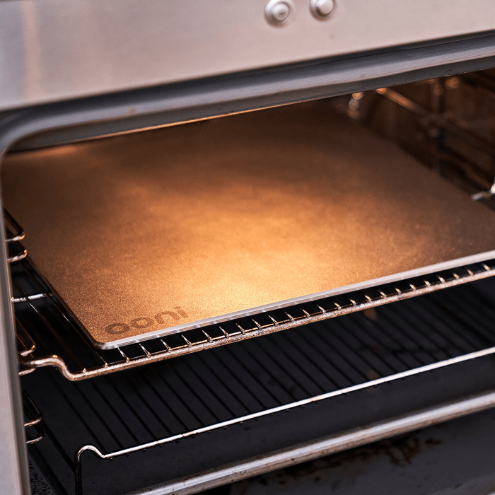 A baking stone rests inside an oven, glowing with heat, reflecting warm light in a metal interior with grates below it.