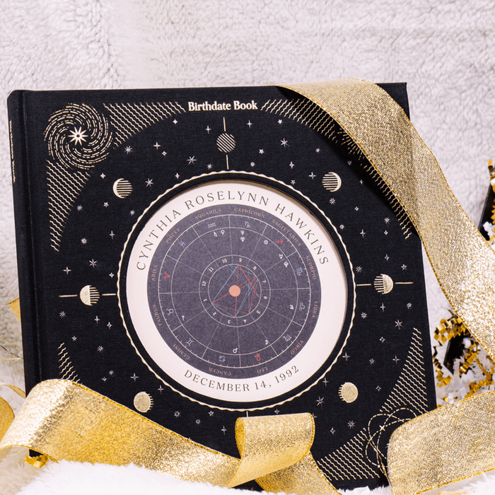 A black book with gold accents displays a birth chart and the name “Cynthia Roselynn Hawkins,” surrounded by gold ribbon on a plush white surface.