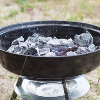 A charcoal grill contains glowing coals, preparing for cooking in an outdoor sandy area surrounded by sparse green grass.