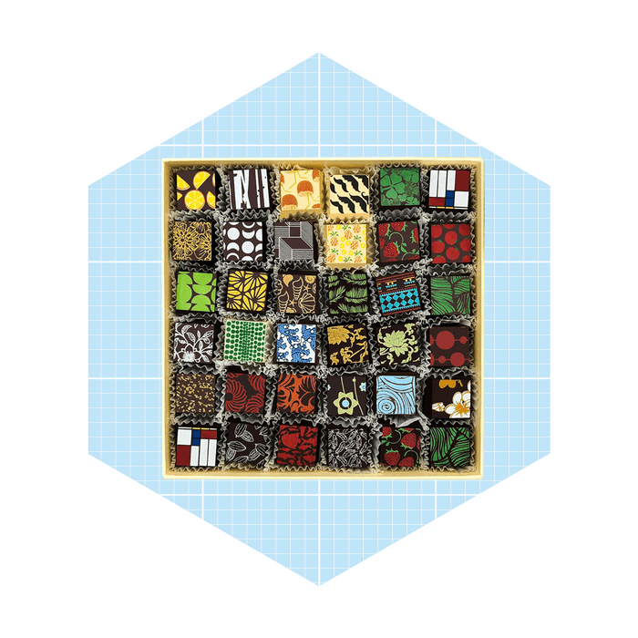A square box displays assorted chocolates, each uniquely patterned and colorful, arranged neatly on a soft paper lining against a light blue grid background.