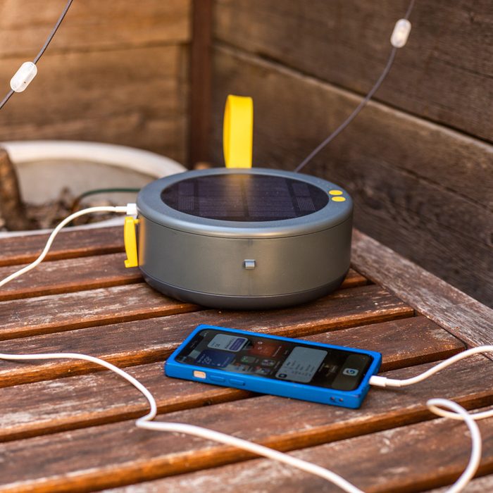 A solar-powered charger sits on a wooden table, charging a smartphone with white cables, surrounded by a rustic wooden backdrop.
