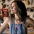 A woman in a striped apron stands confidently, smiling and looking up, surrounded by tools hanging on a workshop wall.