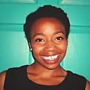 A smiling individual with short, natural hair poses in front of a turquoise wall, exuding warmth and friendliness.