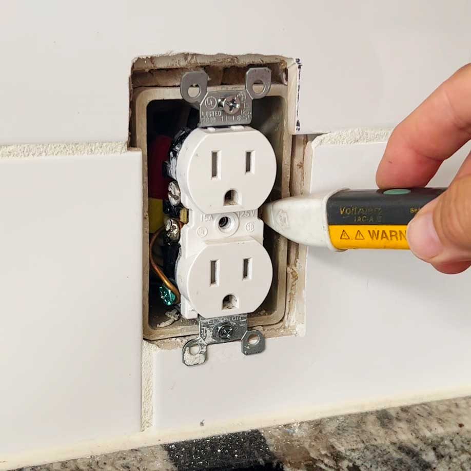A hand uses a voltage tester on an electrical outlet in a wall, revealing internal wiring and components against a granite countertop background.