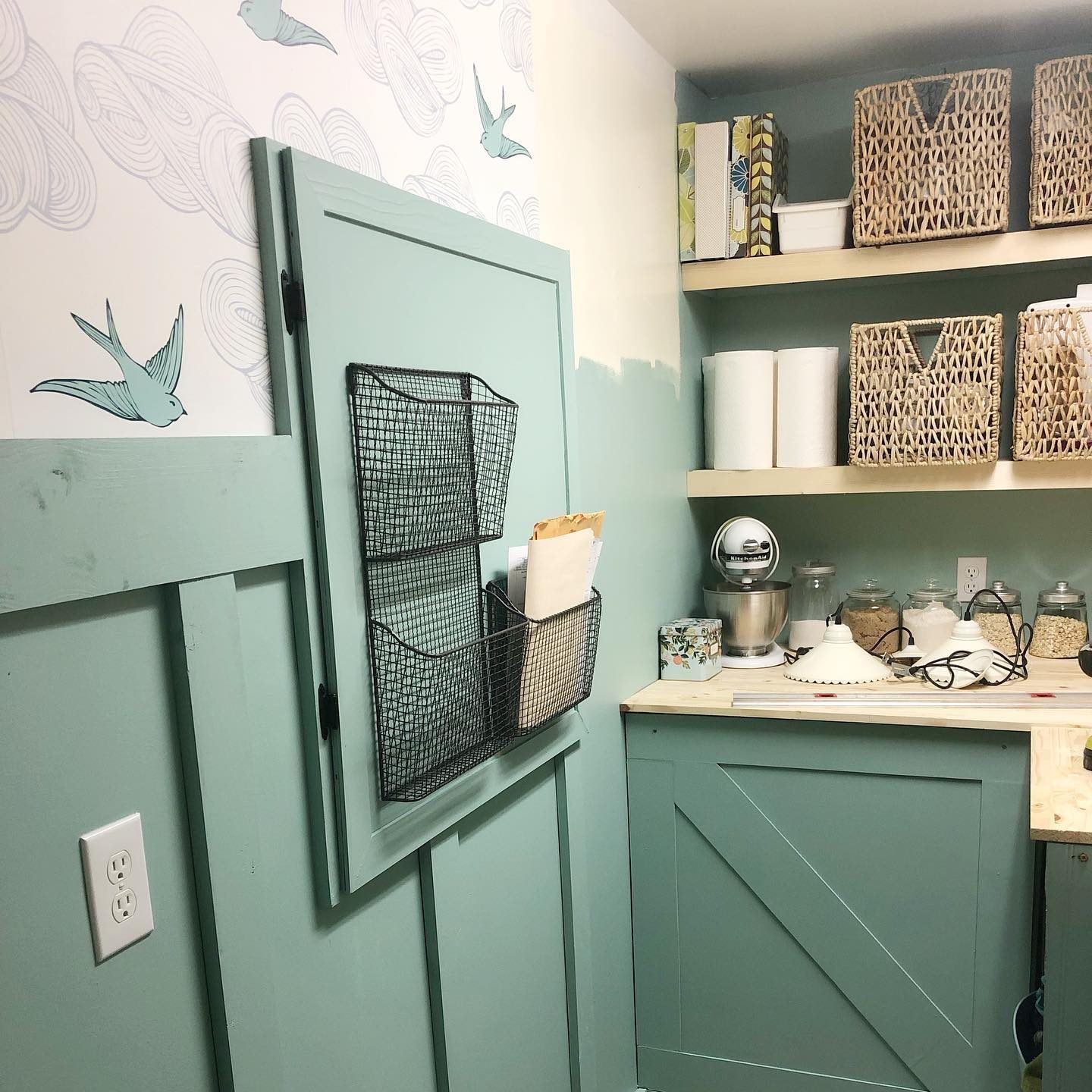 A teal wall with a black wire rack holds papers. A shelving unit displays items, while patterned wallpaper features birds.