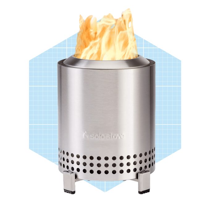 A stainless steel stove emits bright flames from its top, positioned centrally on a blue grid-patterned background, indicating a functional outdoor heating device.