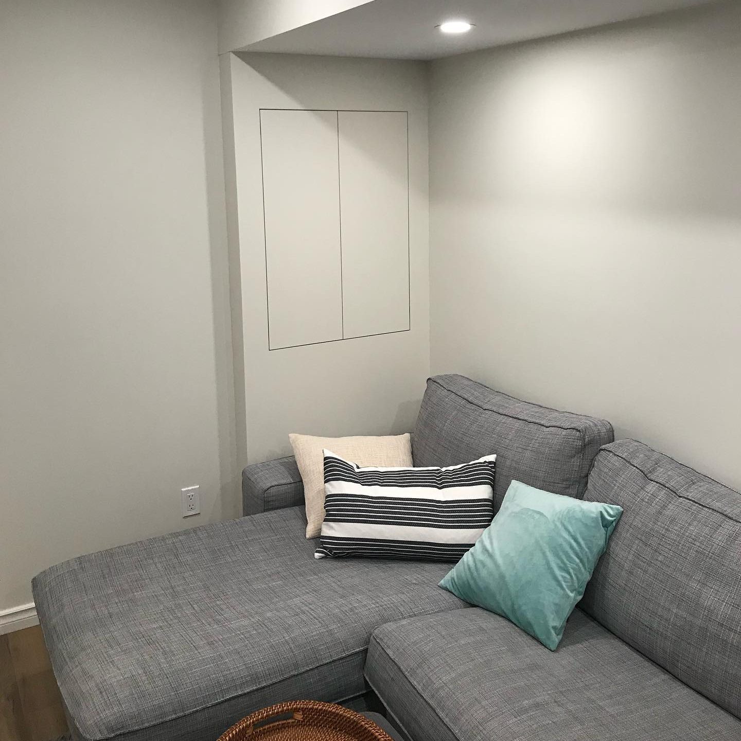 A gray sectional sofa with two decorative pillows reclines in a softly lit, minimalist room with light gray walls and a hidden cabinet.