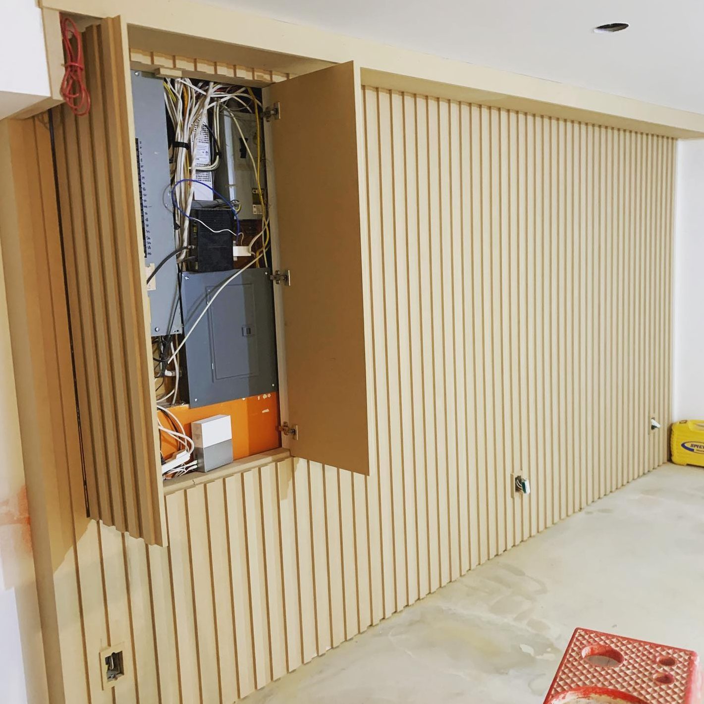 A cabinet with slatted design has its door open, revealing wires and electrical components inside, situated in a partially finished room with bare flooring.