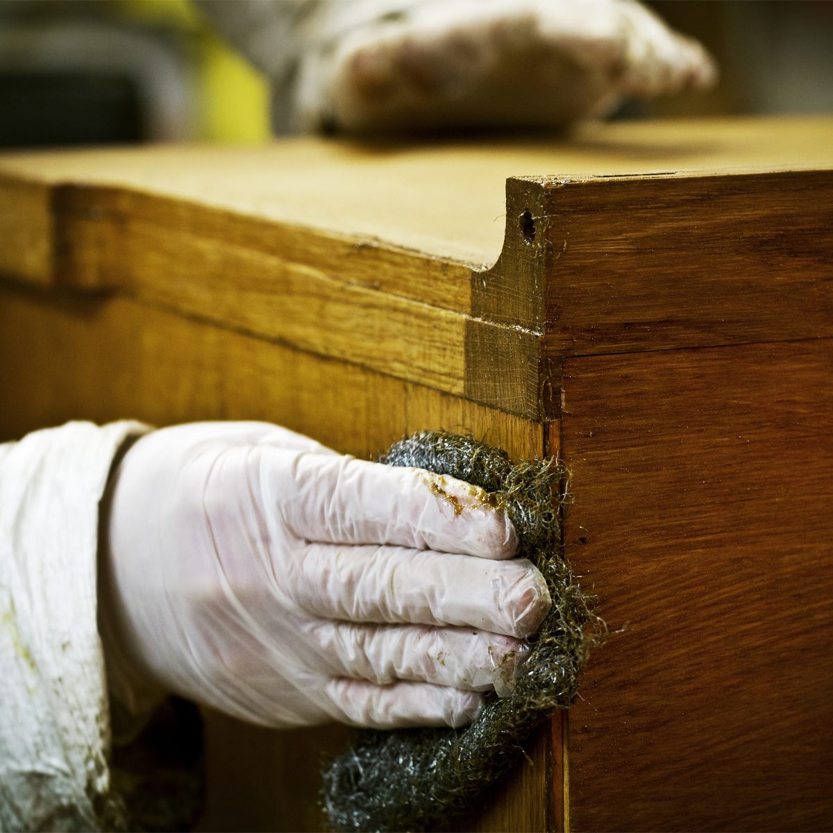 A gloved hand scrubs a wooden furniture edge with a cleaning pad, focusing on restoration in a workshop environment.