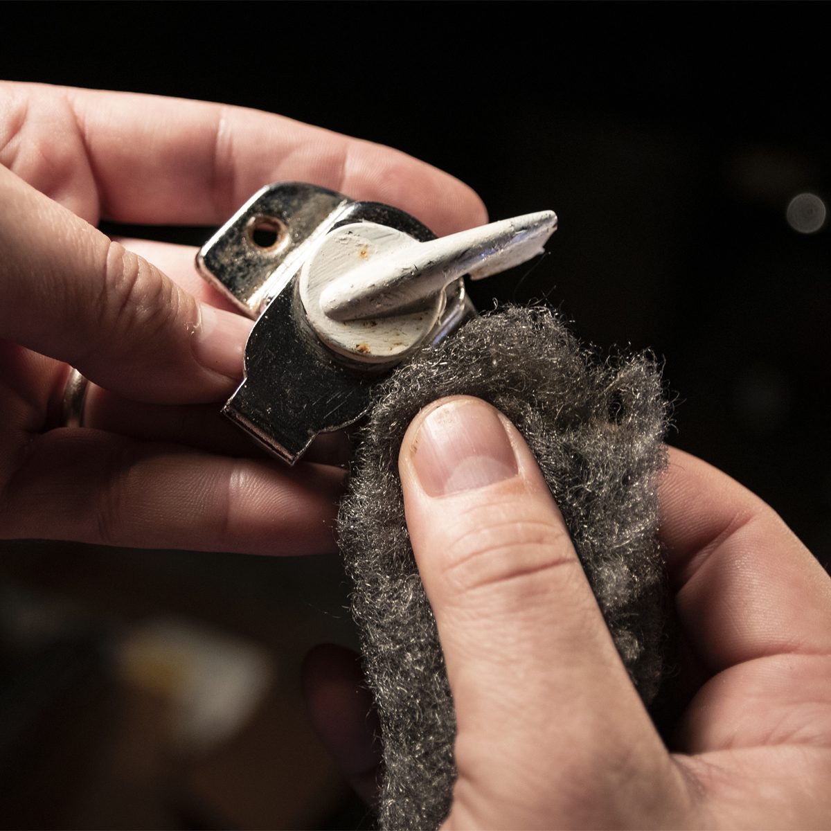 A person cleans a rusty metal object, using a gray cleaning cloth, in a dimly lit space filled with indistinct items.