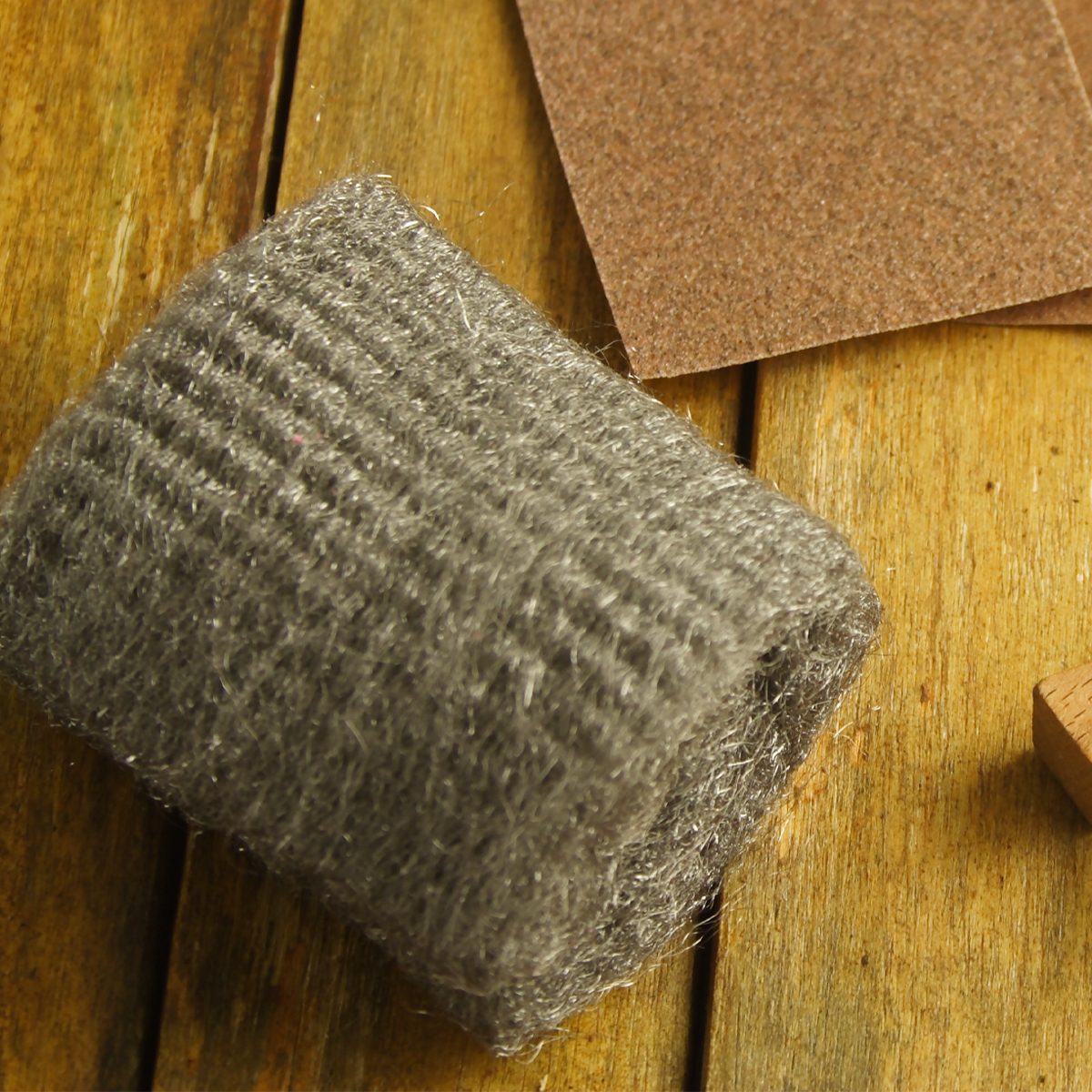 A bundle of gray steel wool sits on a wooden surface with a piece of sandpaper and a small wooden block nearby.