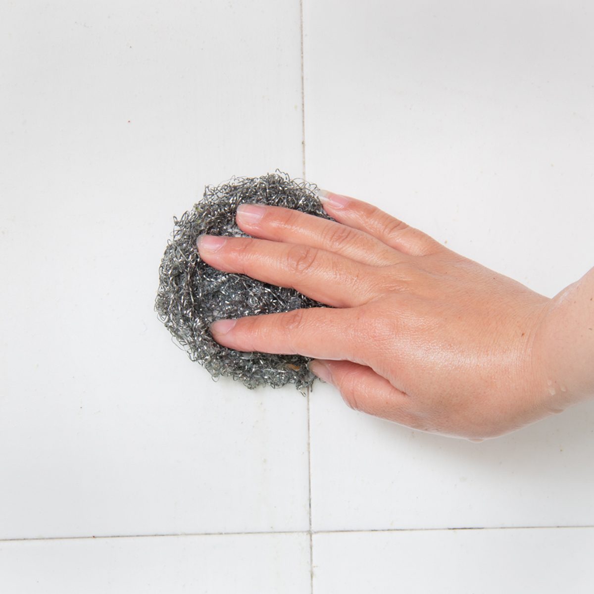 A hand scrubs a shiny metal scouring pad against a white tiled surface, likely cleaning or removing dirt.