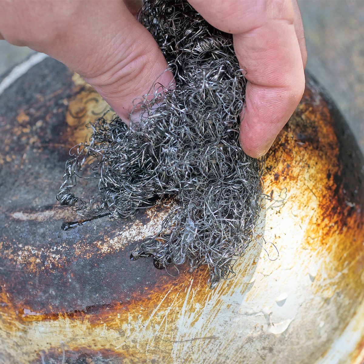 A hand is scrubbing a tarnished metal surface with steel wool, removing grime while the background features a worn and stained bowl or pot.