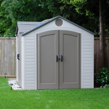 A storage shed stands on green grass, featuring double doors and a gray roof, surrounded by a wooden fence and trees.