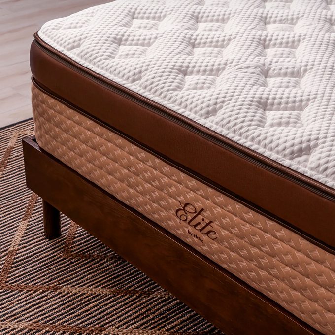 A mattress rests on a wooden frame, featuring a textured surface and a brown border. It's set against a patterned rug in a well-lit room.