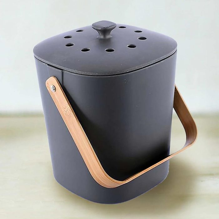 A black kitchen compost bin sits upright, equipped with a bamboo handle and a vented lid, designed for collecting organic waste in a home environment.
