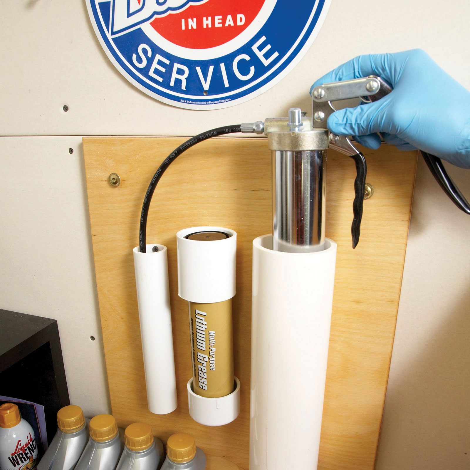 A hand in a blue glove operates a grease dispenser mounted on a wooden wall, near tubes of grease, amid a workshop environment.
