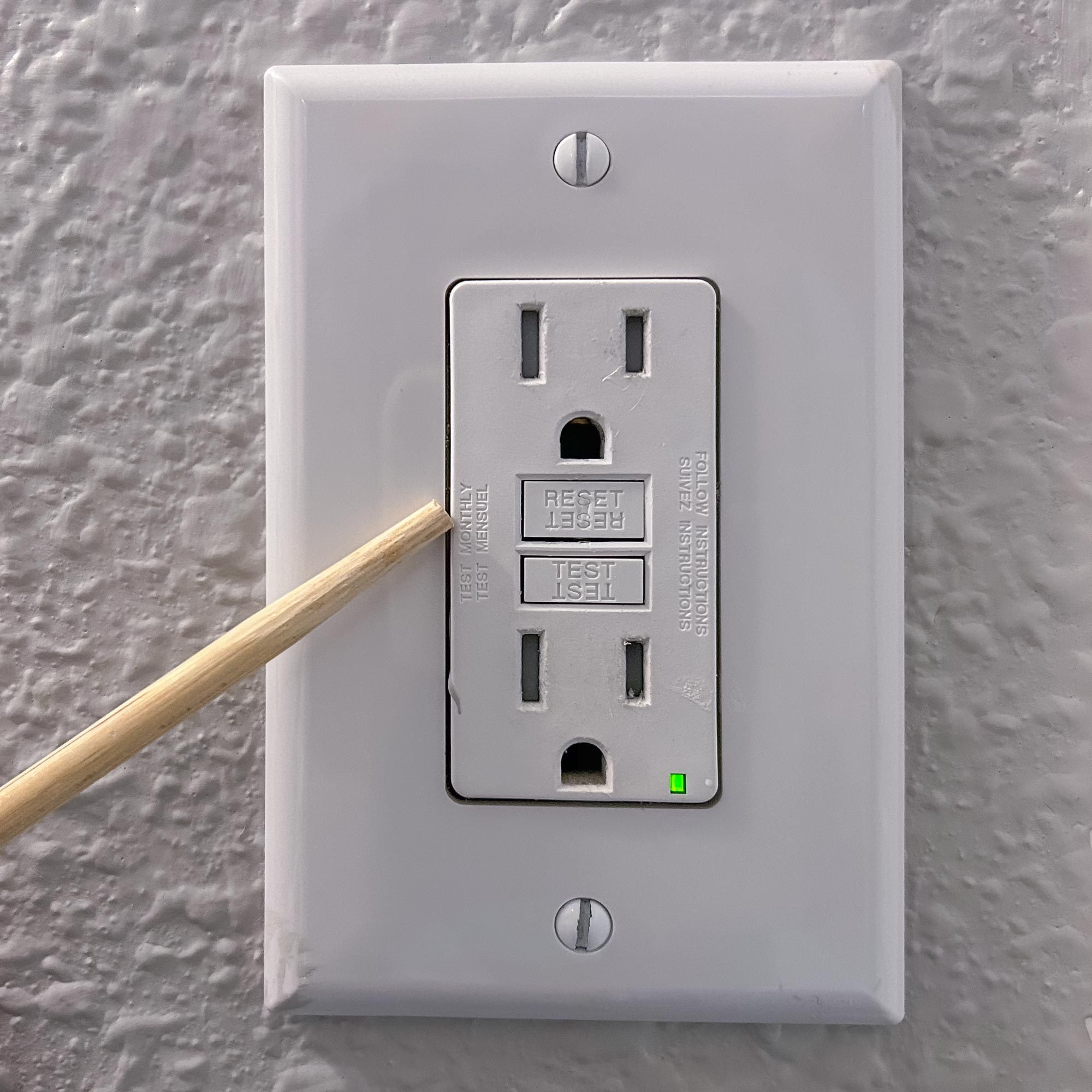 A wooden stick is inserted into a GFCI outlet, which displays a green light, against a textured wall. Buttons labeled "RESET" and "TEST" are visible.
