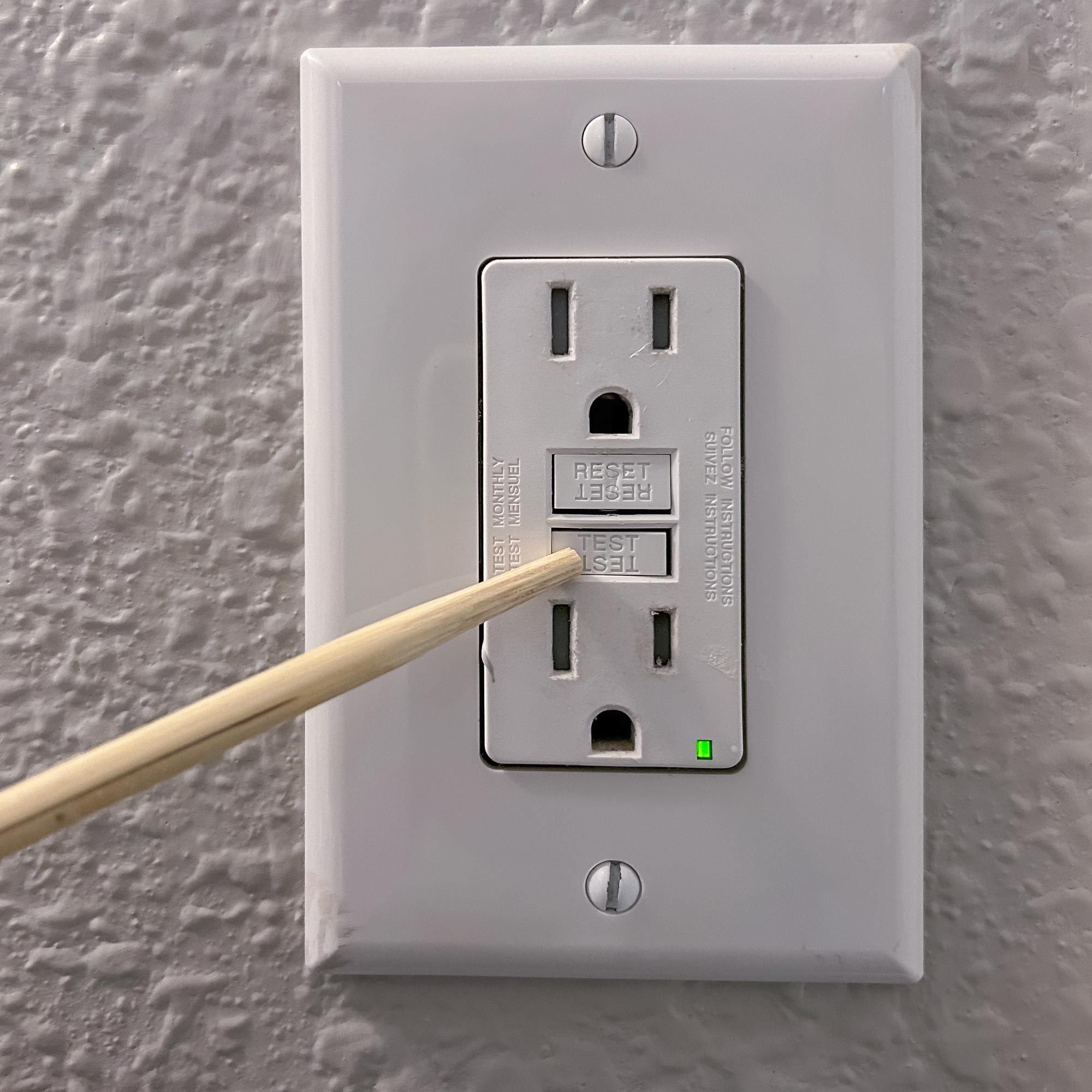A test tool is applied to the "TEST" button on a white electrical outlet, situated on a textured gray wall, showing a green indicator light.