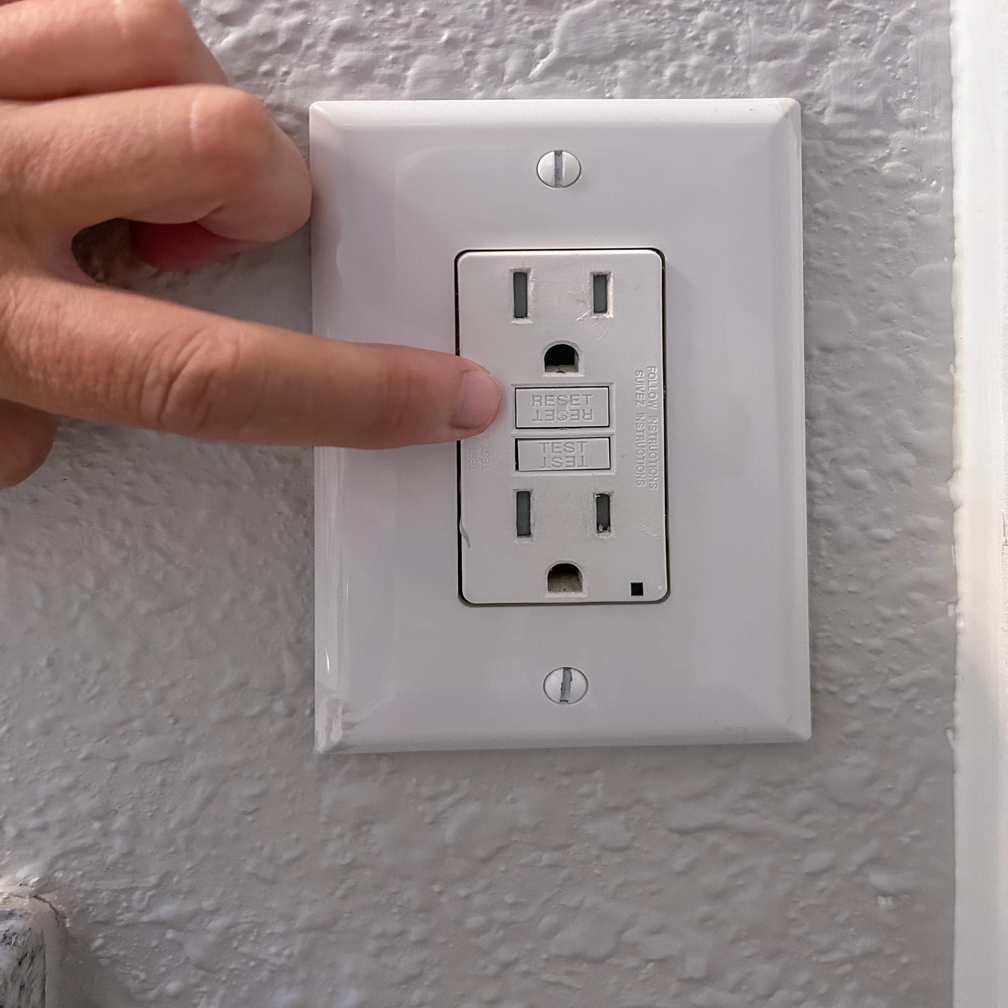A hand presses the "RESET" button on a GFCI outlet mounted on a textured wall, indicating a potential electrical reset procedure.