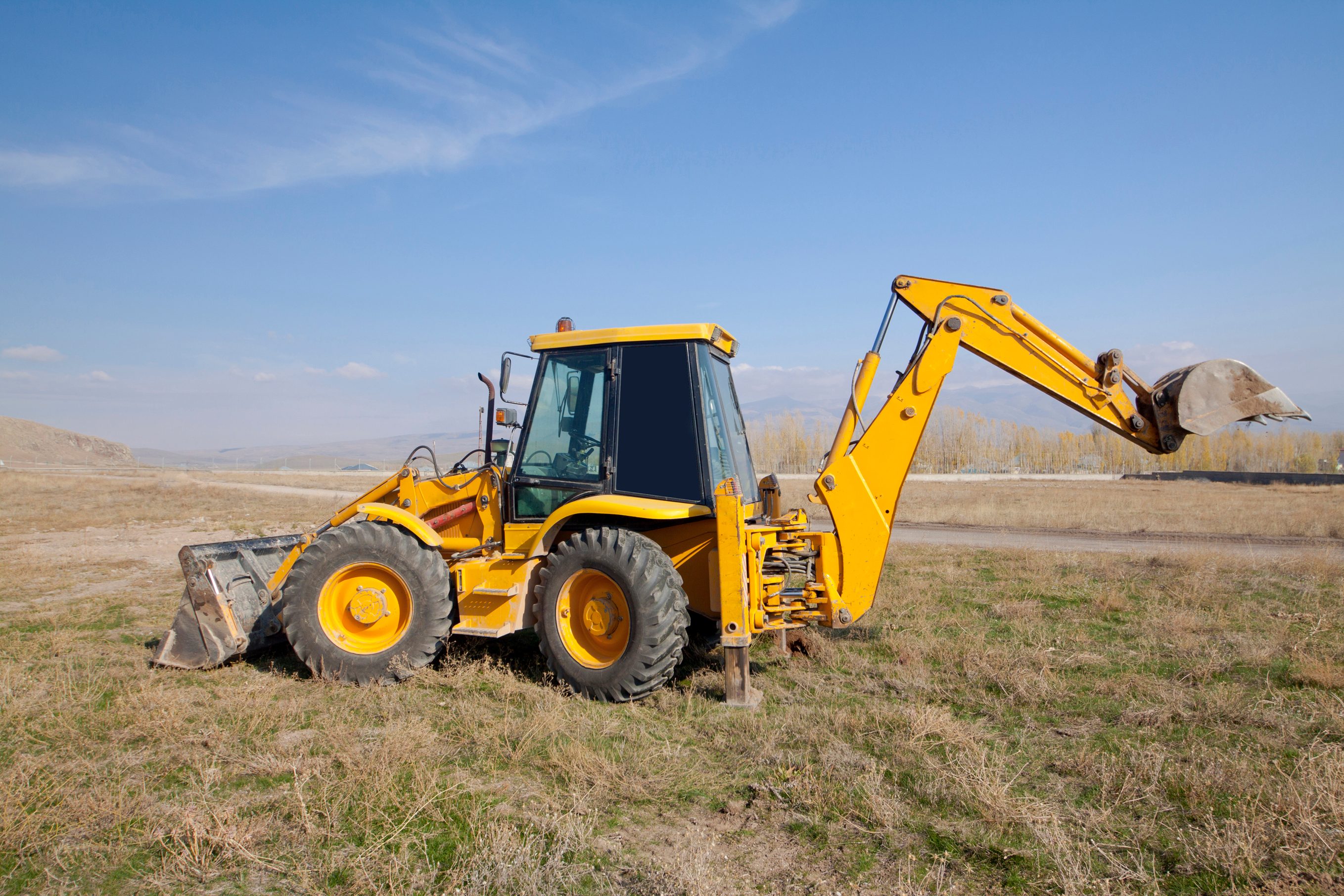 A yellow backhoe sits on grassy terrain, its front bucket lowered, ready to dig, under a clear blue sky with distant hills.