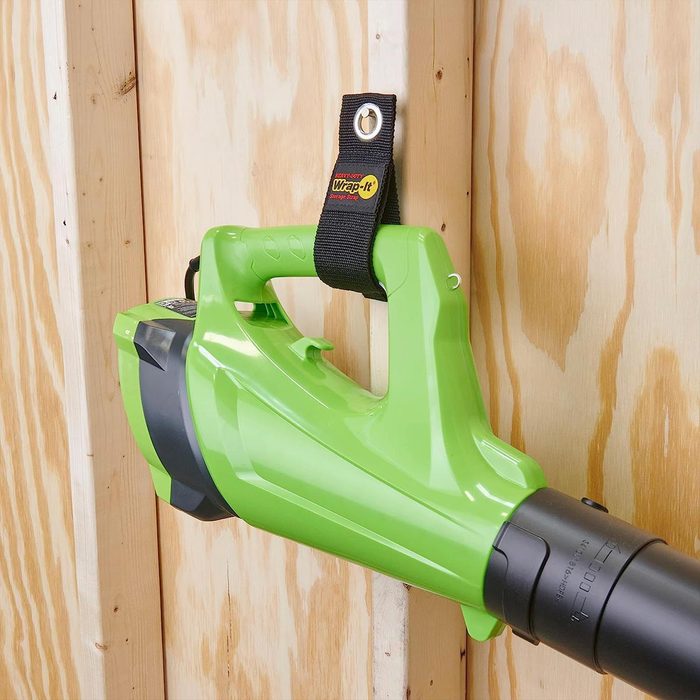A green leaf blower is hung against a wooden wall using a strap, showcasing its design and storage configuration.
