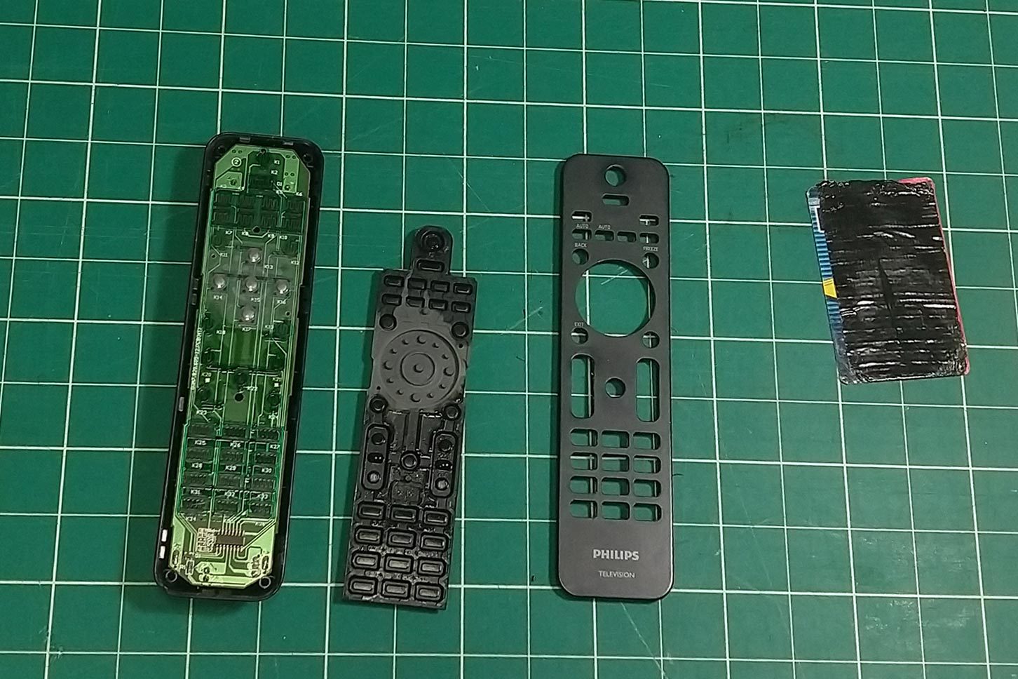 A disassembled remote control lies on a green grid surface, with its circuit board, rubber button pad, plastic casing, and a small card nearby.